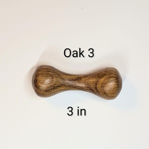 May include: A wooden dumbbell-shaped object made of oak. The object is brown with visible wood grain and has the text "Oak 3" and "3 in" printed on the surface. The dumbbell is 3 inches long.