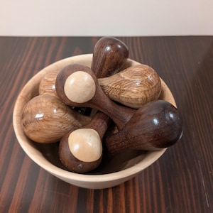 May include: A collection of wooden spoons in a light-colored wooden bowl. The spoons are various shades of brown, with some featuring a lighter, circular accent on the spoon head. The bowl sits on a dark wood surface.
