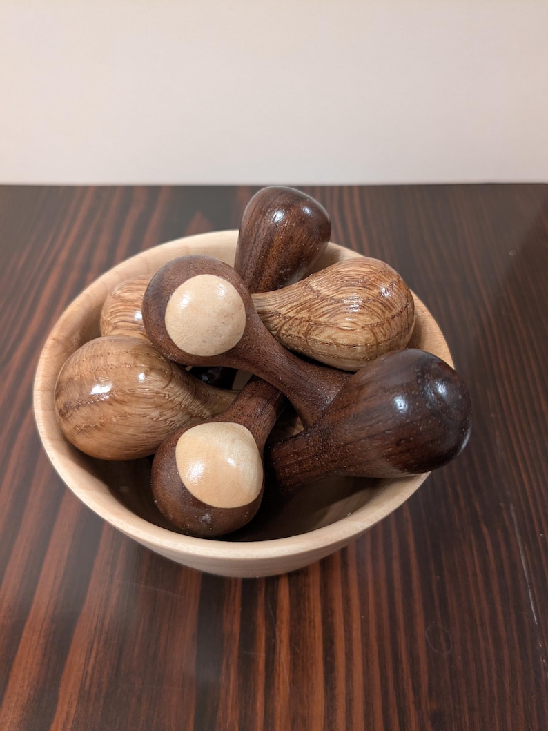 May include: A collection of wooden spoons in a light-colored wooden bowl. The spoons are various shades of brown, with some featuring a lighter, circular accent on the spoon head. The bowl sits on a dark wood surface.