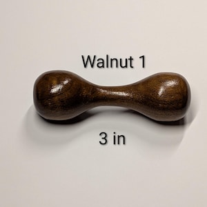 May include: A dumbbell-shaped wooden object, 3 inches long, with a dark brown finish. The text "Walnut 1" is above the object. The dumbbell has rounded ends and a narrower center.