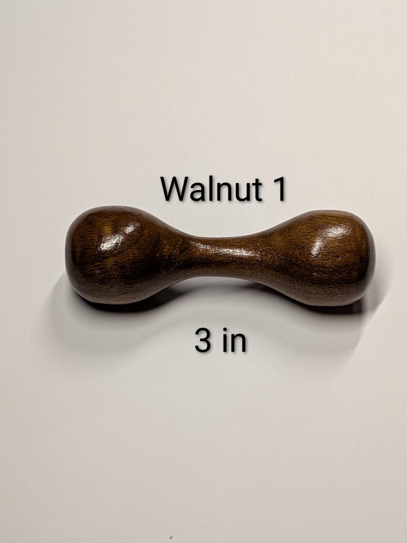 May include: A dumbbell-shaped wooden object, 3 inches long, with a dark brown finish. The text "Walnut 1" is above the object. The dumbbell has rounded ends and a narrower center.