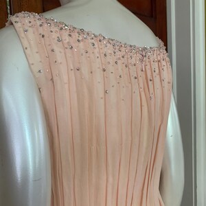 Puede incluir: Un vestido de fiesta de color melocot&oacute;n p&aacute;lido con una falda plisada y un escote con cuentas. El vestido es sin mangas y parece estar hecho de una tela ligera. El escote est&aacute; adornado con peque&ntilde;as cuentas brillantes.