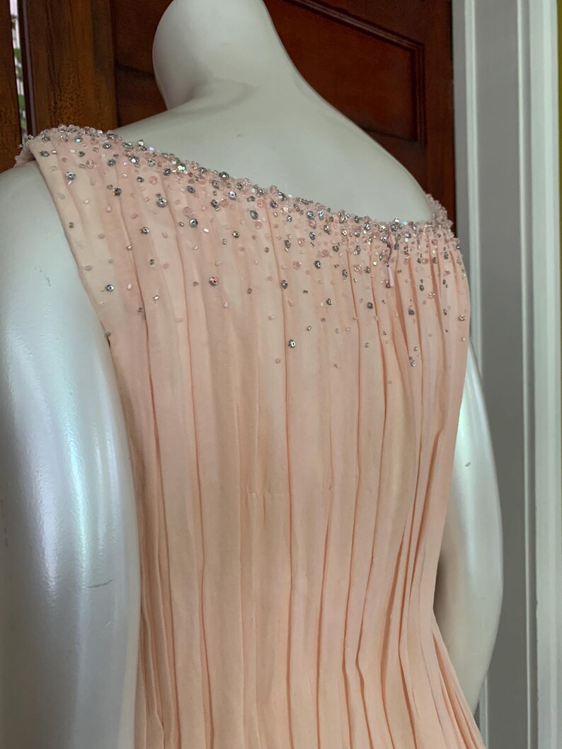 Puede incluir: Un vestido de fiesta de color melocot&oacute;n p&aacute;lido con una falda plisada y un escote con cuentas. El vestido es sin mangas y parece estar hecho de una tela ligera. El escote est&aacute; adornado con peque&ntilde;as cuentas brillantes.