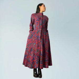 May include: A long, button-down dress with a vibrant paisley pattern in shades of red, blue, and purple. The dress features long sleeves, a high collar, and pockets. The model is wearing black boots.