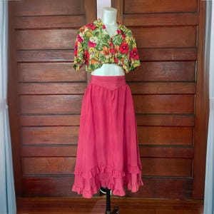 May include: A two-piece outfit displayed on a mannequin. The top is a short-sleeved shirt with a floral pattern in red, yellow, and green. The skirt is a long, flowing, solid pink with a ruffled hem.