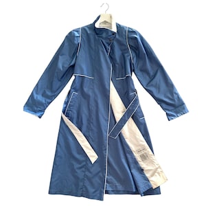 May include: A blue trench coat with white accents, hanging on a hanger. The coat features a wrap design, a belt, and white trim along the edges. The interior lining is white. The coat is made of a shiny material.