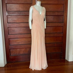 Puede incluir: Un vestido de noche melocot&oacute;n p&aacute;lido sobre un maniqu&iacute;. El vestido presenta un escote en V con adornos de cuentas y un corpi&ntilde;o plisado que fluye hacia una falda larga. El vestido se exhibe frente a una puerta de madera oscura.