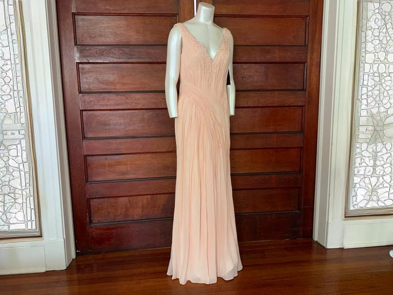 Puede incluir: Un vestido de noche melocot&oacute;n p&aacute;lido sobre un maniqu&iacute;. El vestido presenta un escote en V con adornos de cuentas y un corpi&ntilde;o plisado que fluye hacia una falda larga. El vestido se exhibe frente a una puerta de madera oscura.