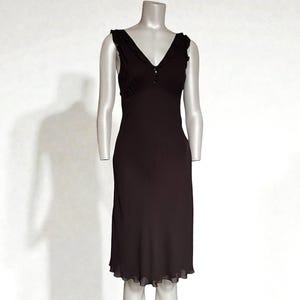 May include: A dark brown sleeveless dress with a V-neckline and a knee-length hem. The dress features delicate ruffle detailing along the neckline and a subtle gathering at the bust. The dress is displayed on a mannequin.