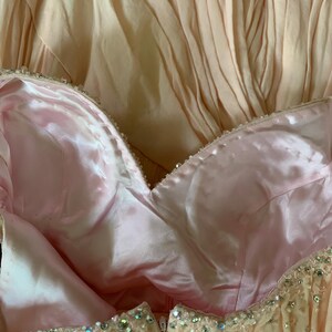 Puede incluir: Primer plano de un vestido formal en tonos rosa p&aacute;lido y melocot&oacute;n. El corpi&ntilde;o presenta un escote coraz&oacute;n con forro de sat&eacute;n y est&aacute; adornado con cuentas brillantes. La falda tiene un dise&ntilde;o plisado.