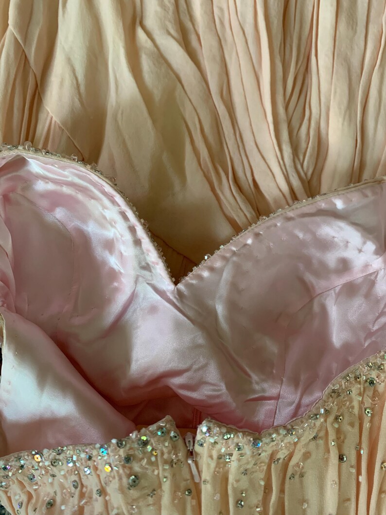 Puede incluir: Primer plano de un vestido formal en tonos rosa p&aacute;lido y melocot&oacute;n. El corpi&ntilde;o presenta un escote coraz&oacute;n con forro de sat&eacute;n y est&aacute; adornado con cuentas brillantes. La falda tiene un dise&ntilde;o plisado.