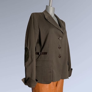 Könnte beinhalten: Ein brauner Nadelstreifen-Blazer mit fallendem Revers, drei Knöpfen und einem kleinen Gürteldetail. Der Blazer hat Ellbogenpatches und wird auf einer Schaufensterpuppe präsentiert. Die Manschetten sind hochgekrempelt.