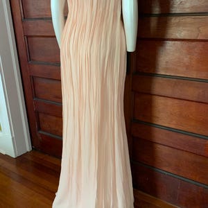 Puede incluir: Un vestido largo de color melocot&oacute;n p&aacute;lido con pliegues verticales. El vestido es sin mangas y parece estar hecho de una tela fluida y ligera. El vestido se exhibe en un maniqu&iacute;.
