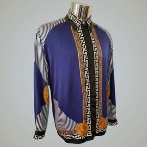 以下が含まれることがあります： 印象的なデザインの長袖シャツ。 シャツは、ネイビーブルーのボディ、白と黒のストライプの袖、黒の襟が特徴です。 金と白の幾何学模様とメダリオンが前面を飾っています。