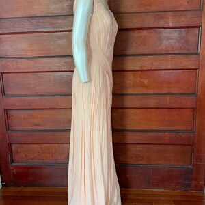 Puede incluir: Un vestido de noche melocot&oacute;n p&aacute;lido sin mangas. El vestido presenta delicados detalles de cuentas en el corpi&ntilde;o y una falda plisada fluida. El vestido se exhibe en un maniqu&iacute;, mostrando su elegante silueta.