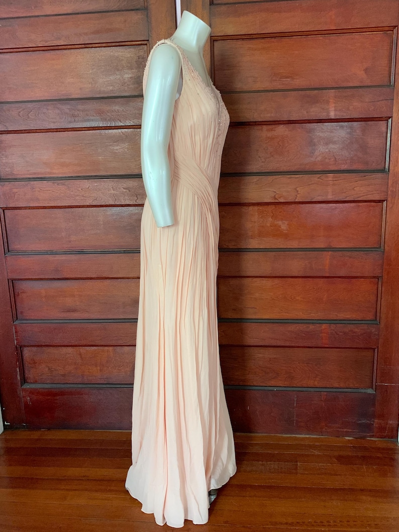 Puede incluir: Un vestido de noche melocot&oacute;n p&aacute;lido sin mangas. El vestido presenta delicados detalles de cuentas en el corpi&ntilde;o y una falda plisada fluida. El vestido se exhibe en un maniqu&iacute;, mostrando su elegante silueta.