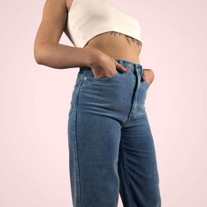 May include: A white crop top paired with high-waisted, wide-leg corduroy pants in a light blue hue. The pants feature a classic five-pocket design and a button and zipper closure. The outfit is casual and stylish.