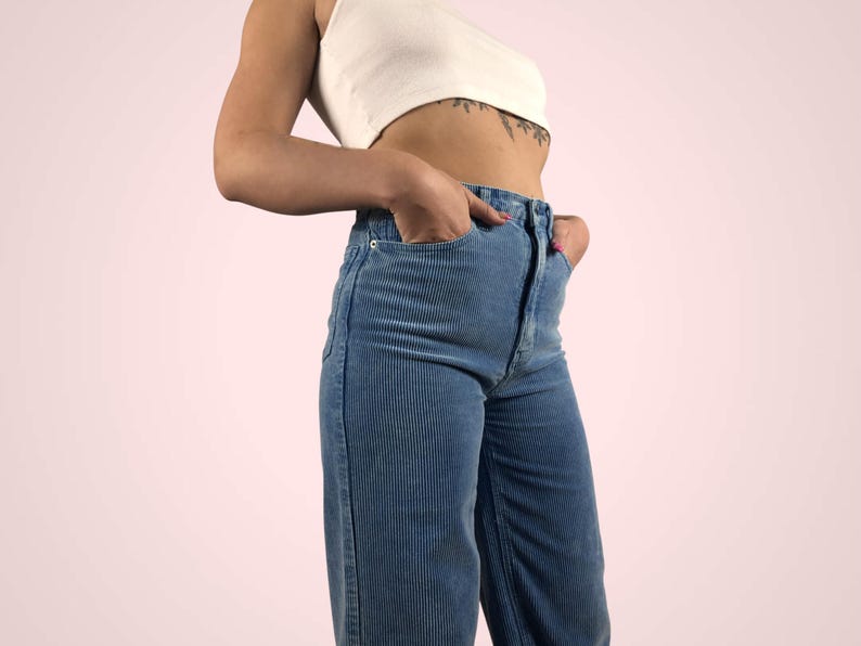May include: A white crop top paired with high-waisted, wide-leg corduroy pants in a light blue hue. The pants feature a classic five-pocket design and a button and zipper closure. The outfit is casual and stylish.