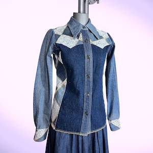May include: A denim shirt and skirt set on a mannequin. The long-sleeved shirt features a collared neckline, button-down front, and decorative white lace and gold trim. The skirt is pleated. The outfit is a mix of light and dark blue denim.