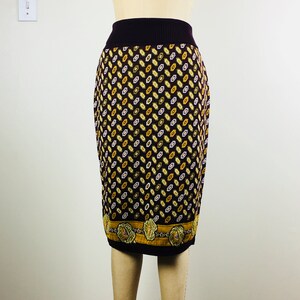May include: A knee-length skirt with a dark brown background and a repeating pattern of yellow, white, and gold diamond shapes. The skirt has a ribbed, dark purple waistband and a decorative border at the hem. The skirt is displayed on a mannequin.