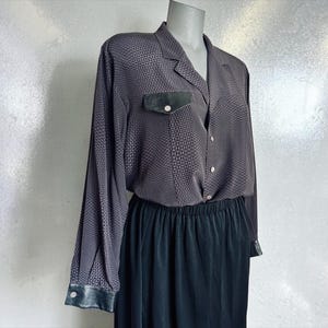 May include: A long-sleeved button-down shirt in a dark gray and black geometric pattern, featuring a notched collar, a single chest pocket with a black flap, and black cuffs. The shirt is paired with a solid black skirt with an elastic waistband.
