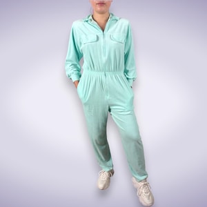 Op de afbeelding: Licht turquoise jumpsuit met een kraag, lange mouwen en een ritssluiting. De jumpsuit heeft twee borstzakken, een elastische taille en rechte pijpen. De persoon draagt witte sneakers.