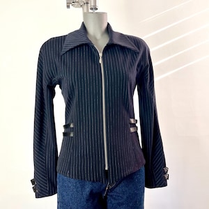 May include: A dark navy blue long-sleeve jacket featuring a white pinstripe pattern, a large pointed collar, and a full-length silver front zipper. The jacket includes two black horizontal straps at each side of the waist and at the cuffs.