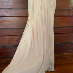 Puede incluir: Un vestido largo color melocot&oacute;n p&aacute;lido con un corpi&ntilde;o plisado y una falda fluida. La tela se drapea elegantemente, creando pliegues suaves y un brillo sutil. El vestido se exhibe en un maniqu&iacute;.
