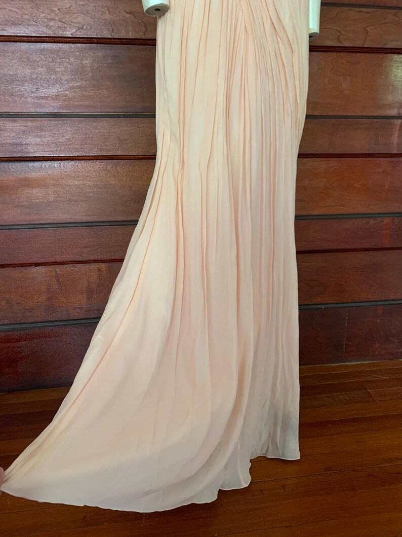 Puede incluir: Un vestido largo color melocot&oacute;n p&aacute;lido con un corpi&ntilde;o plisado y una falda fluida. La tela se drapea elegantemente, creando pliegues suaves y un brillo sutil. El vestido se exhibe en un maniqu&iacute;.