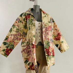 Puede incluir: Un abrigo con estampado floral con una base de color crema y un vibrante patrón de flores rojas, rosas y moradas. El abrigo tiene una parte delantera abotonada, mangas largas y un cuello. El abrigo se exhibe en un maniquí.