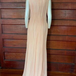 Puede incluir: Un vestido de noche melocot&oacute;n p&aacute;lido con un corpi&ntilde;o plisado y una falda fluida. El vestido sin mangas presenta un escote con cuentas y se exhibe en un maniqu&iacute;. El vestido largo es adecuado para ocasiones especiales.