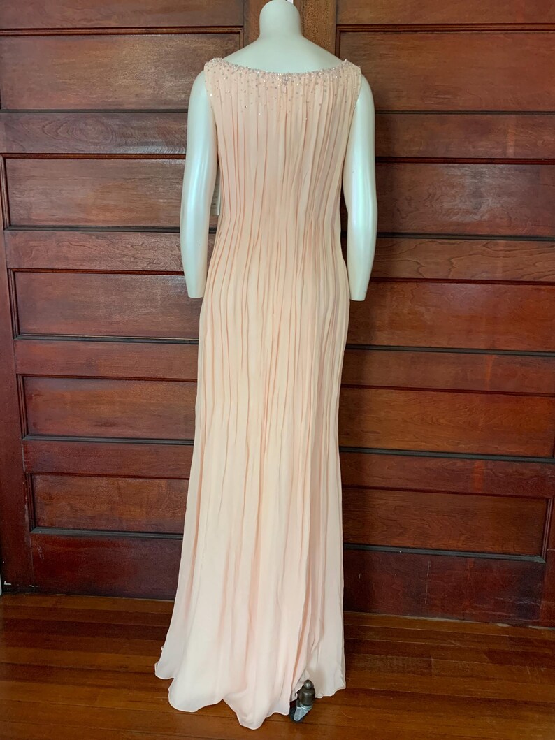 Puede incluir: Un vestido de noche melocot&oacute;n p&aacute;lido con un corpi&ntilde;o plisado y una falda fluida. El vestido sin mangas presenta un escote con cuentas y se exhibe en un maniqu&iacute;. El vestido largo es adecuado para ocasiones especiales.