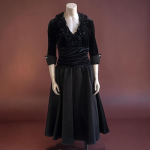 May include: A black vintage dress on a mannequin. The dress features a velvet bodice with a ruffled neckline and three-quarter sleeves. The waist is cinched with a velvet band, and the skirt is a flowing, full length design.