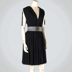 Puede incluir: Vestido negro con cuello en V y falda a la rodilla. El vestido se ciñe a la cintura con un cinturón plateado con una hebilla decorativa. El vestido tiene mangas cortas con correas negras. El maniquí está sobre un fondo blanco.