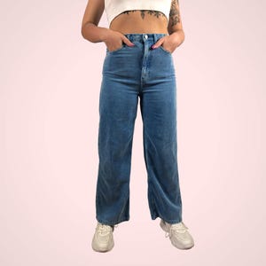 May include: High-waisted, wide-leg blue corduroy pants. The pants have a classic five-pocket design and a button and zipper closure. The model is wearing white sneakers. The pants are a medium wash blue.