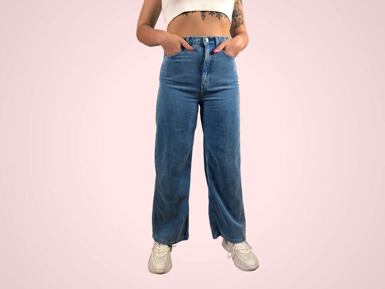 May include: High-waisted, wide-leg blue corduroy pants. The pants have a classic five-pocket design and a button and zipper closure. The model is wearing white sneakers. The pants are a medium wash blue.