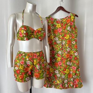 Könnte beinhalten: Ein zweiteiliges Set bestehend aus einem Neckholder-Bikini-Oberteil und passender Shorts sowie einem ärmellosen Kleid mit durchgehender Knopfleiste und gerüschtem Saum. Alle Teile haben einen olivgrünen Hintergrund mit einem floralen Muster in Rot, Orange, Gelb und Weiß.
