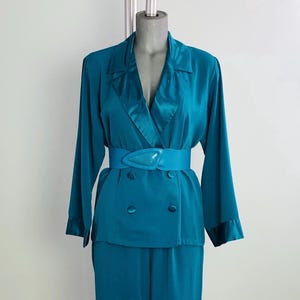 May include: A teal blue satin suit with a double-breasted blazer and a matching skirt. The blazer has a wide collar and a belt with a turquoise buckle.