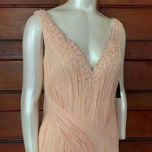 Puede incluir: Un vestido de noche color melocot&oacute;n con escote en V y adornos de cuentas. El vestido presenta un dise&ntilde;o fluido y plisado y se exhibe en un maniqu&iacute;. El vestido es sin mangas y tiene una apariencia suave y elegante.