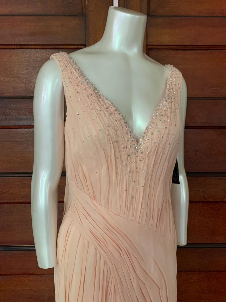 Puede incluir: Un vestido de noche color melocot&oacute;n con escote en V y adornos de cuentas. El vestido presenta un dise&ntilde;o fluido y plisado y se exhibe en un maniqu&iacute;. El vestido es sin mangas y tiene una apariencia suave y elegante.