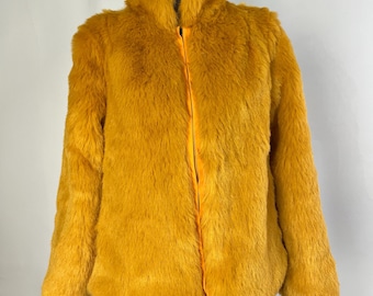 May include: A mustard yellow faux fur jacket featuring a high collar and a central vertical zipper closure. The garment is crafted from a thick, textured synthetic material with long, soft fibers, providing a uniform golden-yellow color throughout.