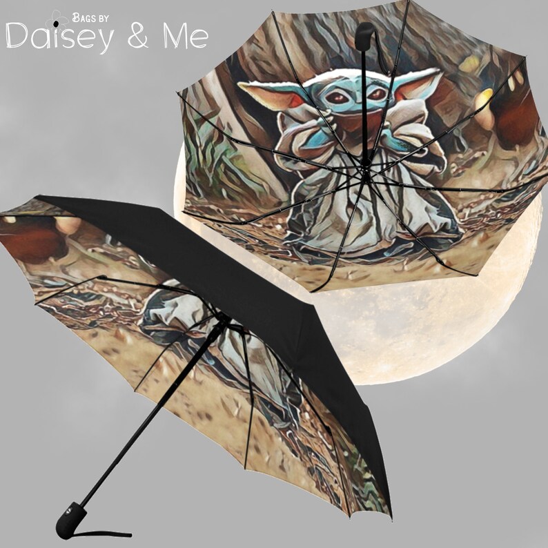May include: A black umbrella with a colorful image of a character from the Star Wars franchise, known as "The Child" or "Baby Yoda", standing in a forest setting. The umbrella is open and the character is facing the viewer. The umbrella is against a gray background.