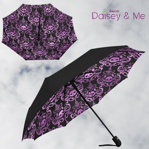 Può includere: Un ombrello nero con un motivo damascato rosa e nero con teschi. L'ombrello è aperto e il manico è visibile. Il testo "Bags by Daisey & Me" è visibile nell'angolo in alto a destra dell'immagine.