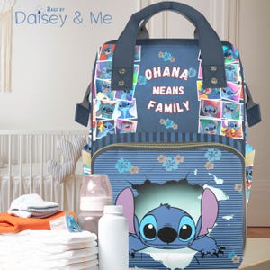 May include: A navy blue diaper bag featuring Stitch from Lilo & Stitch. The bag has a top handle and multiple compartments. The front of the bag has the words "OHANA MEANS FAMILY" and a graphic of Stitch. The bag is surrounded by baby supplies.
