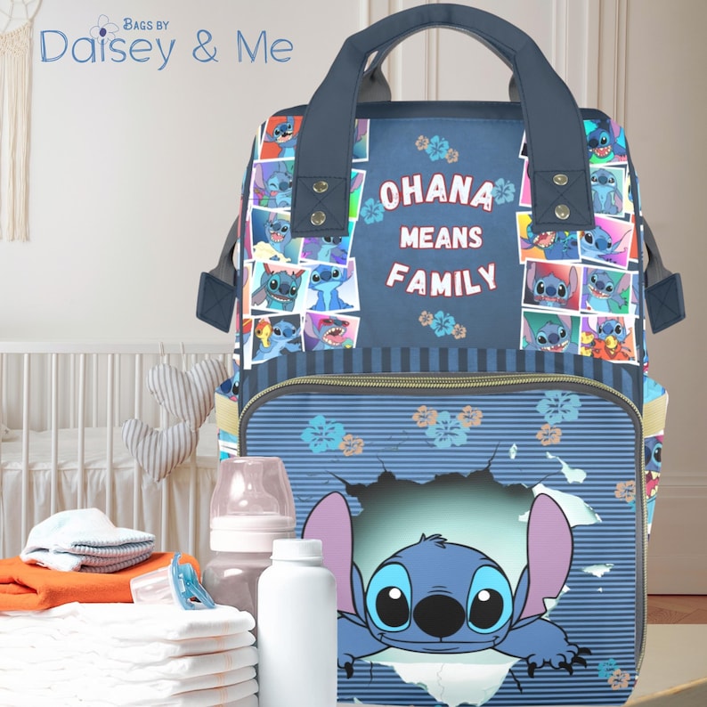 May include: A navy blue diaper bag featuring Stitch from Lilo & Stitch. The bag has a top handle and multiple compartments. The front of the bag has the words "OHANA MEANS FAMILY" and a graphic of Stitch. The bag is surrounded by baby supplies.
