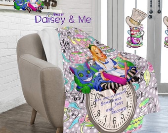 May include: A gray blanket featuring a whimsical Alice in Wonderland design. The blanket showcases Alice, the Cheshire Cat, teacups, and a clock with the text "How long is forever? Sometimes just one second." The blanket is draped over a gray armchair.