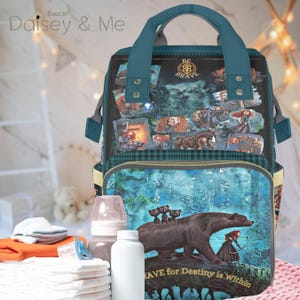 May include: A teal blue diaper bag with a  "Be Brave" design featuring a bear and a girl with a bow and arrow. The bag has a large pocket on the front and a smaller pocket on the top. The bag is sitting on a table with a white surface.