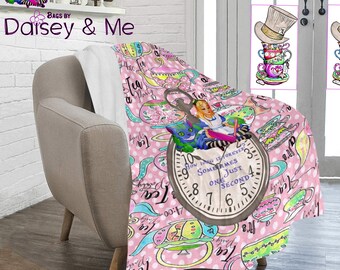 May include: A pink and white fleece throw blanket with a whimsical Alice in Wonderland theme. The blanket features a repeating pattern of teacups, teapots, and clocks. The center of the blanket features Alice and the Cheshire Cat sitting on a pocket watch with the quote "How long is forever? Sometimes just one second."