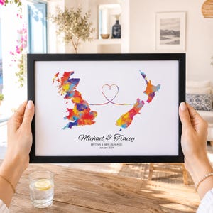 Peut inclure: Une impression encadrée présentant une carte aquarelle colorée de la Grande-Bretagne et de la Nouvelle-Zélande reliées par une ligne fine formant un cœur. Le texte indique Michael & Tracey, BRITAIN & NEW ZEALAND, et January 2024 en noir.