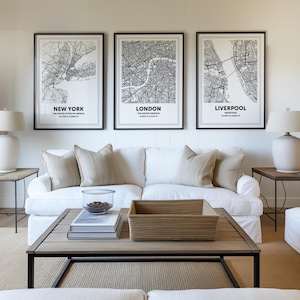 May include: Three framed black and white city map prints of New York, London, and Liverpool hang above a white sofa with beige cushions. A wooden coffee table with a woven basket and books sits in front of the sofa. Two white lamps are on side tables.